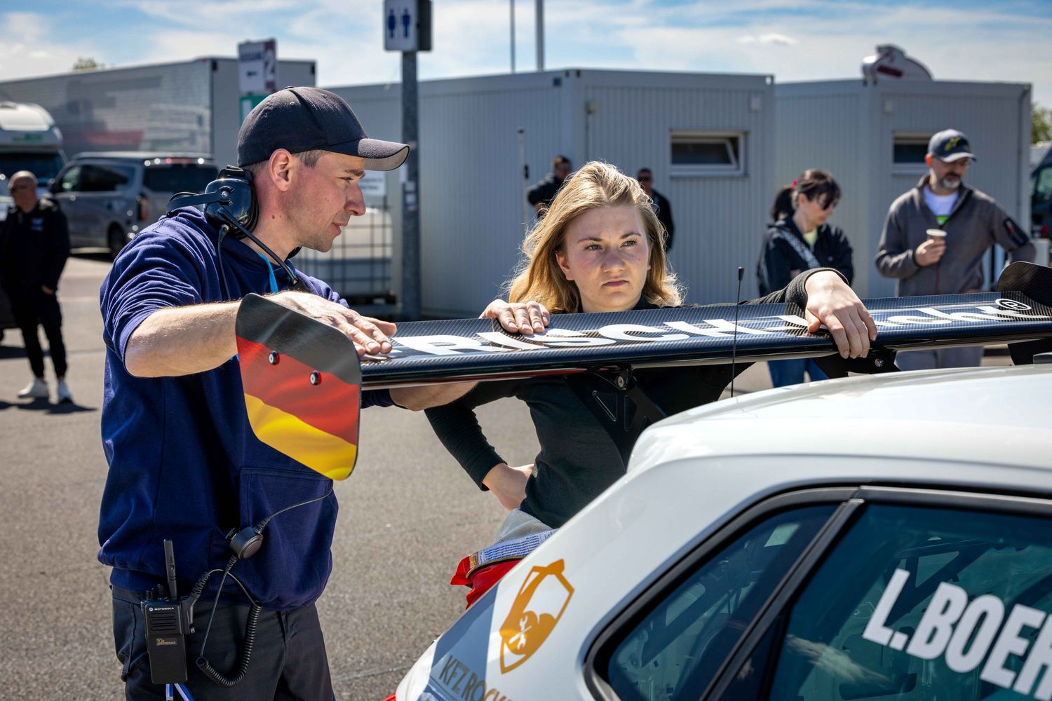 A mechanic and female racing driver inspect a car spoiler at a motorsport event, with a German flag sticker visible.