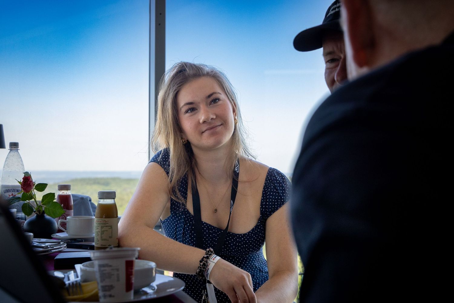Young blonde woman smiling at a table in a bright restaurant with scenic views through large windows.