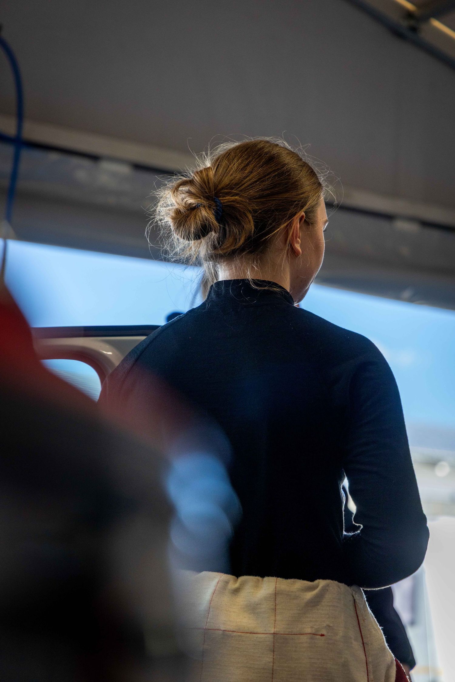Woman with hair bun in dark jacket stands looking out window on a boat or ferry.