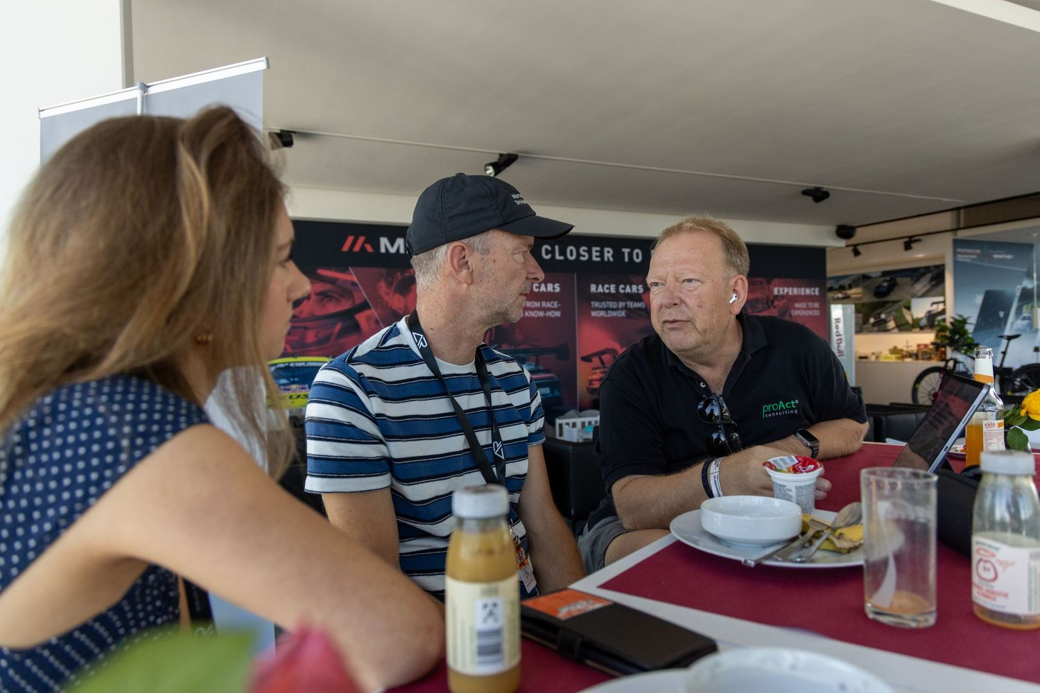 Three people having a conversation at an outdoor dining table with food and drinks at a sailing event.