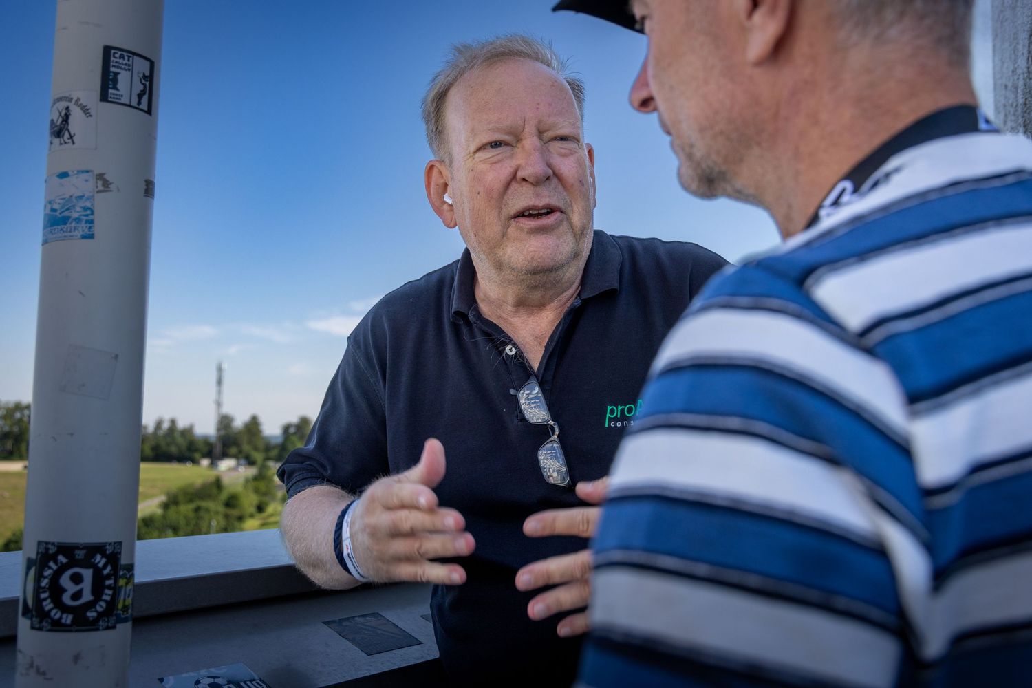 Two men engaged in conversation outdoors on a sunny day, one gesturing expressively in a dark polo shirt.