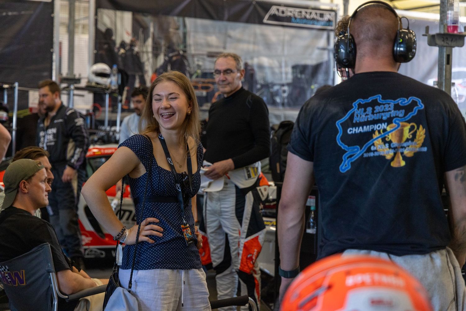 A woman laughs joyfully in a racing garage while a crew member in headphones stands nearby during a motorsport event.