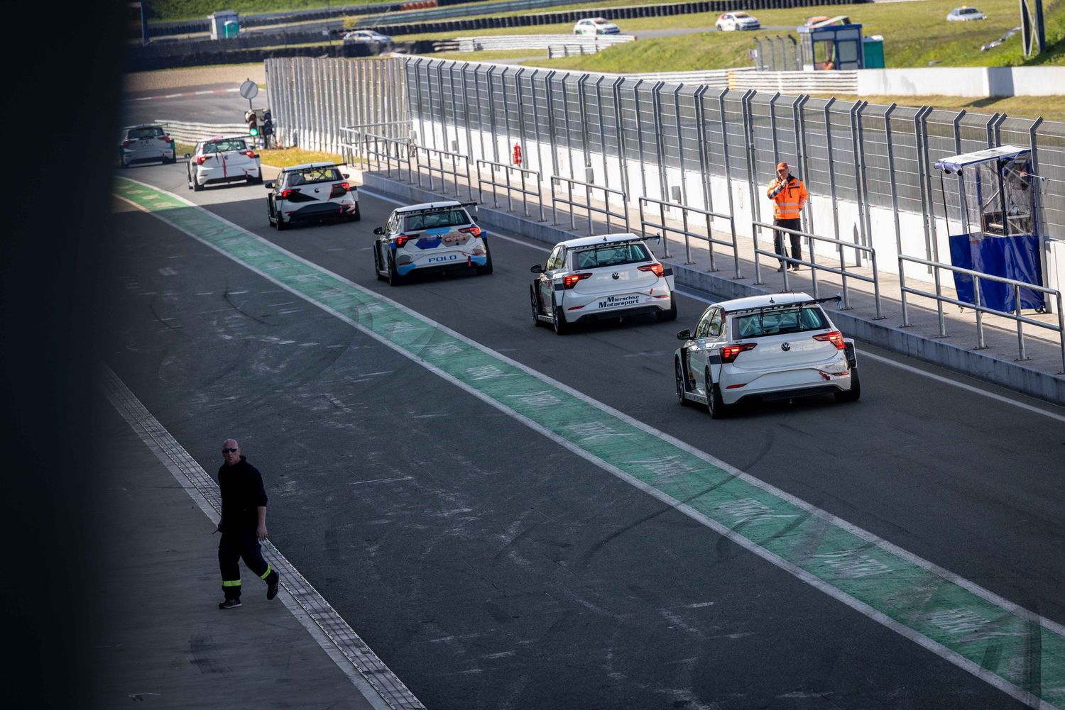 Racing cars line up on a motorsport circuit track with marshals standing by the pit lane barrier.