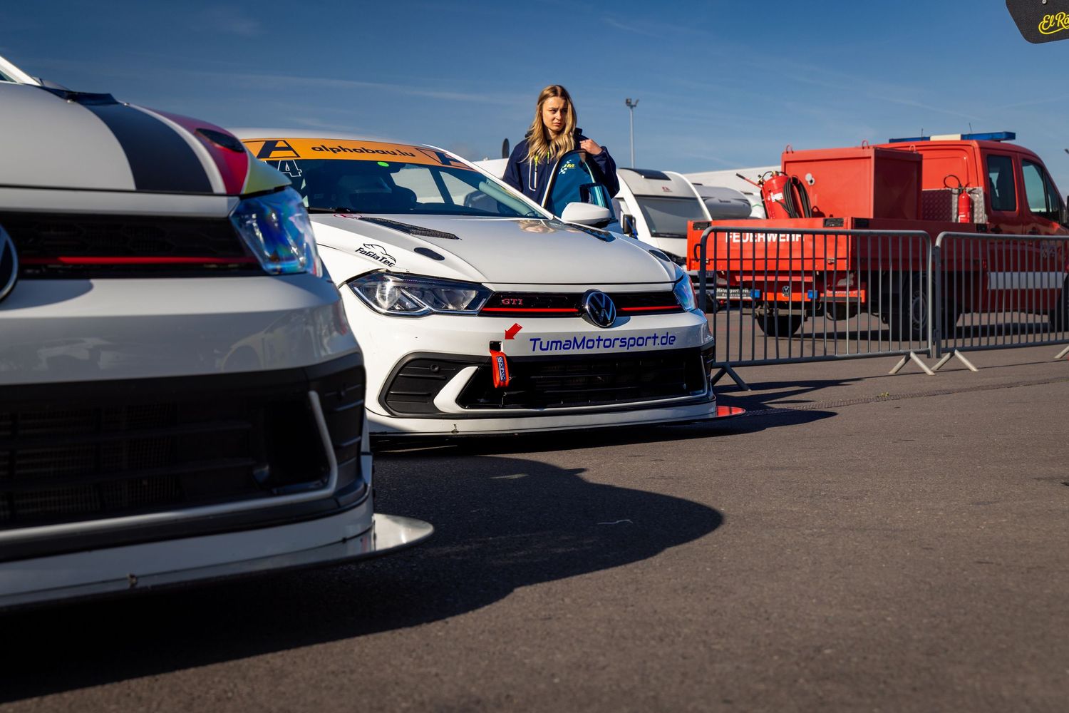 Female racing driver stands beside white Volkswagen race cars in a motorsport paddock on a sunny day.