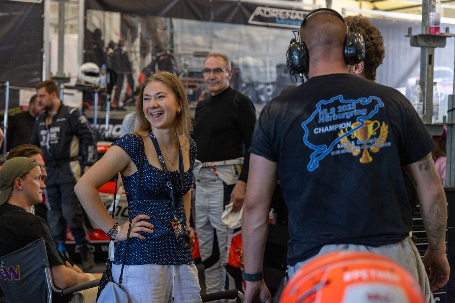 Woman smiling in a racing pit garage surrounded by crew members in a motorsport event setting.