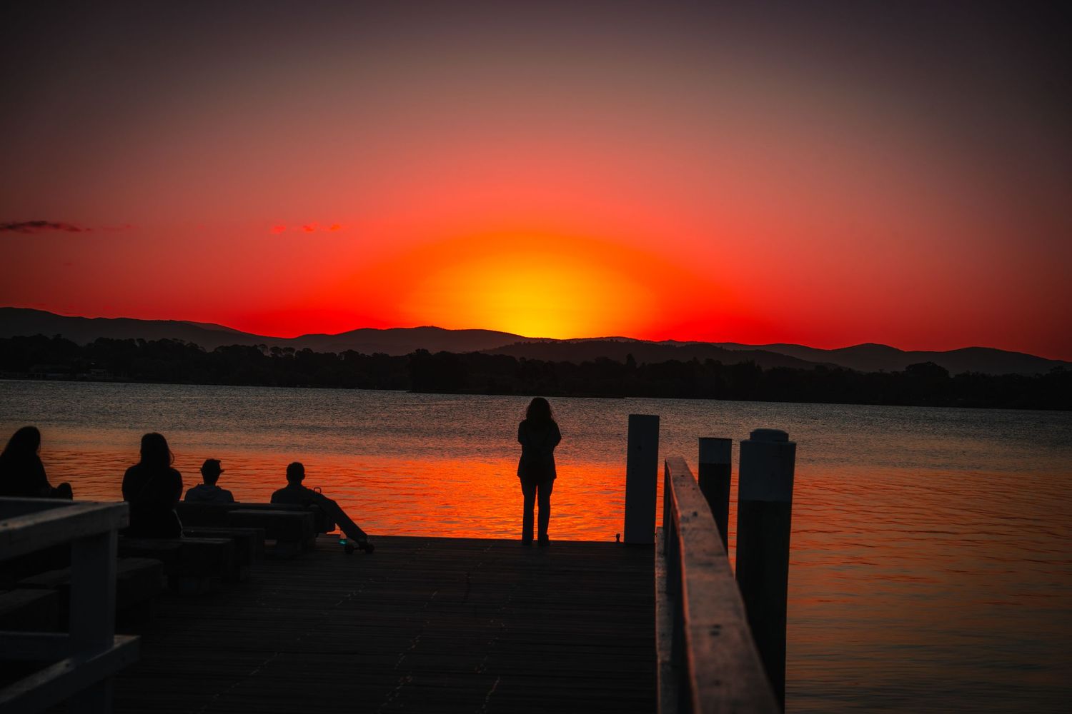 A vibrant red sunset silhouettes figures and posts along a waterfront.