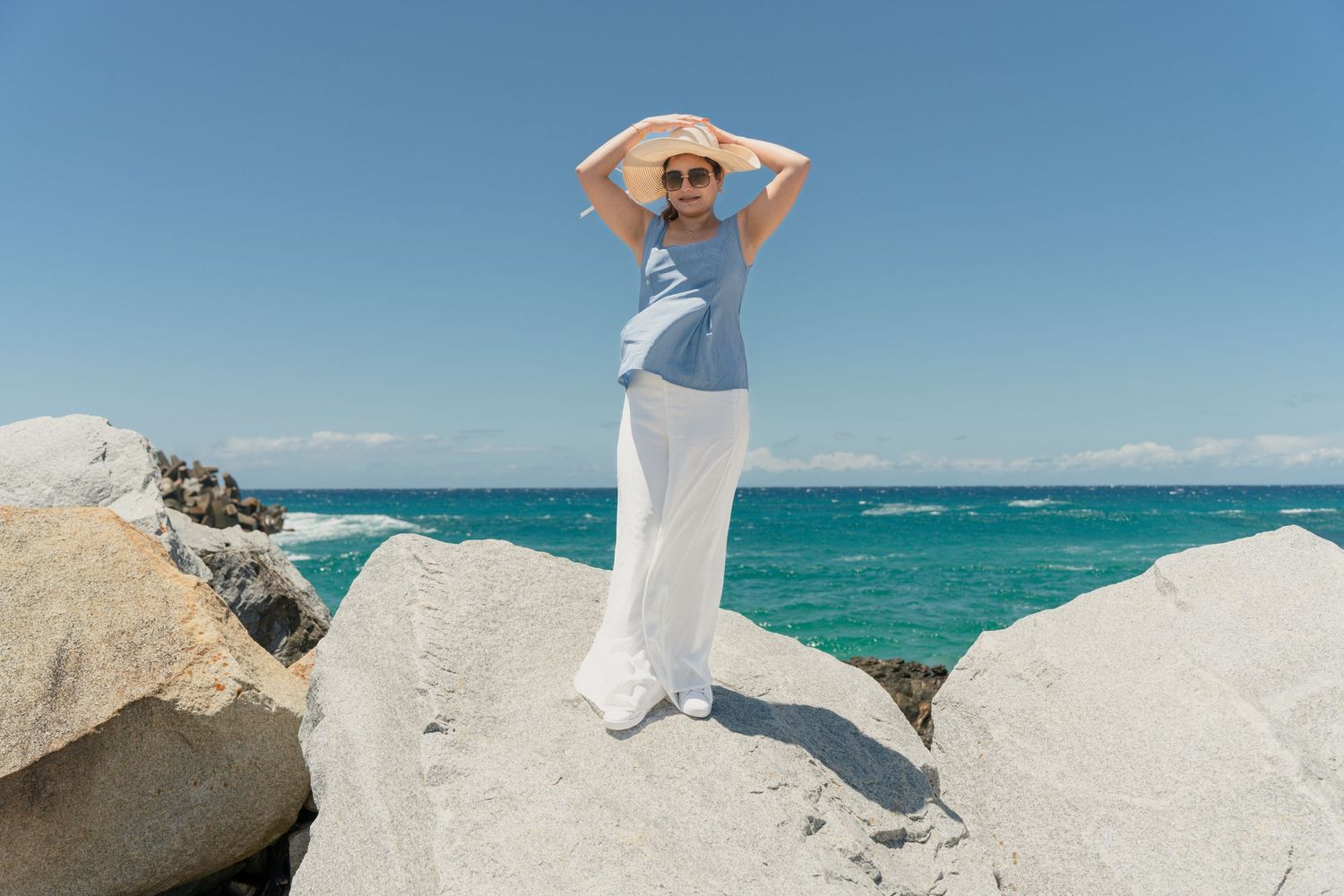 Person in white outfit poses on coastal rocks overlooking bright blue ocean waters.
