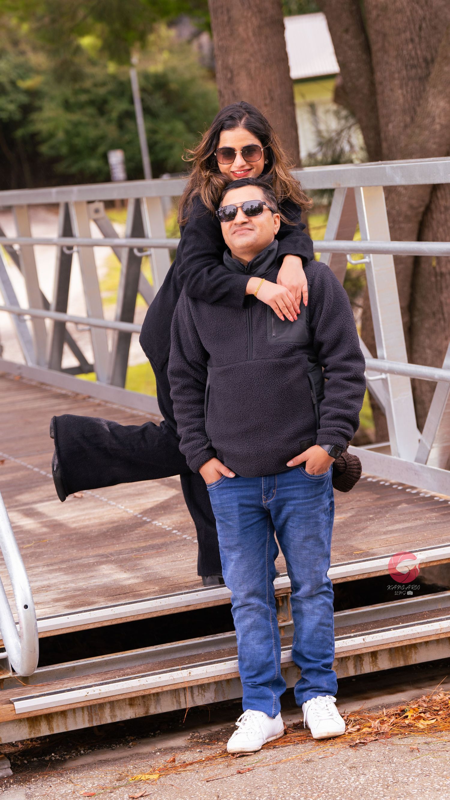 Two people in casual wear share intimate moment on wooden deck with trees in background.