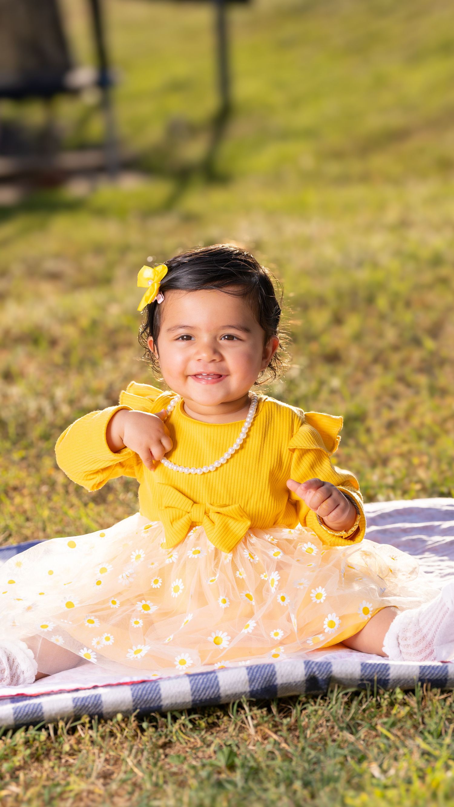 Baby in yellow dress smiling while lying on floral blanket in grass.