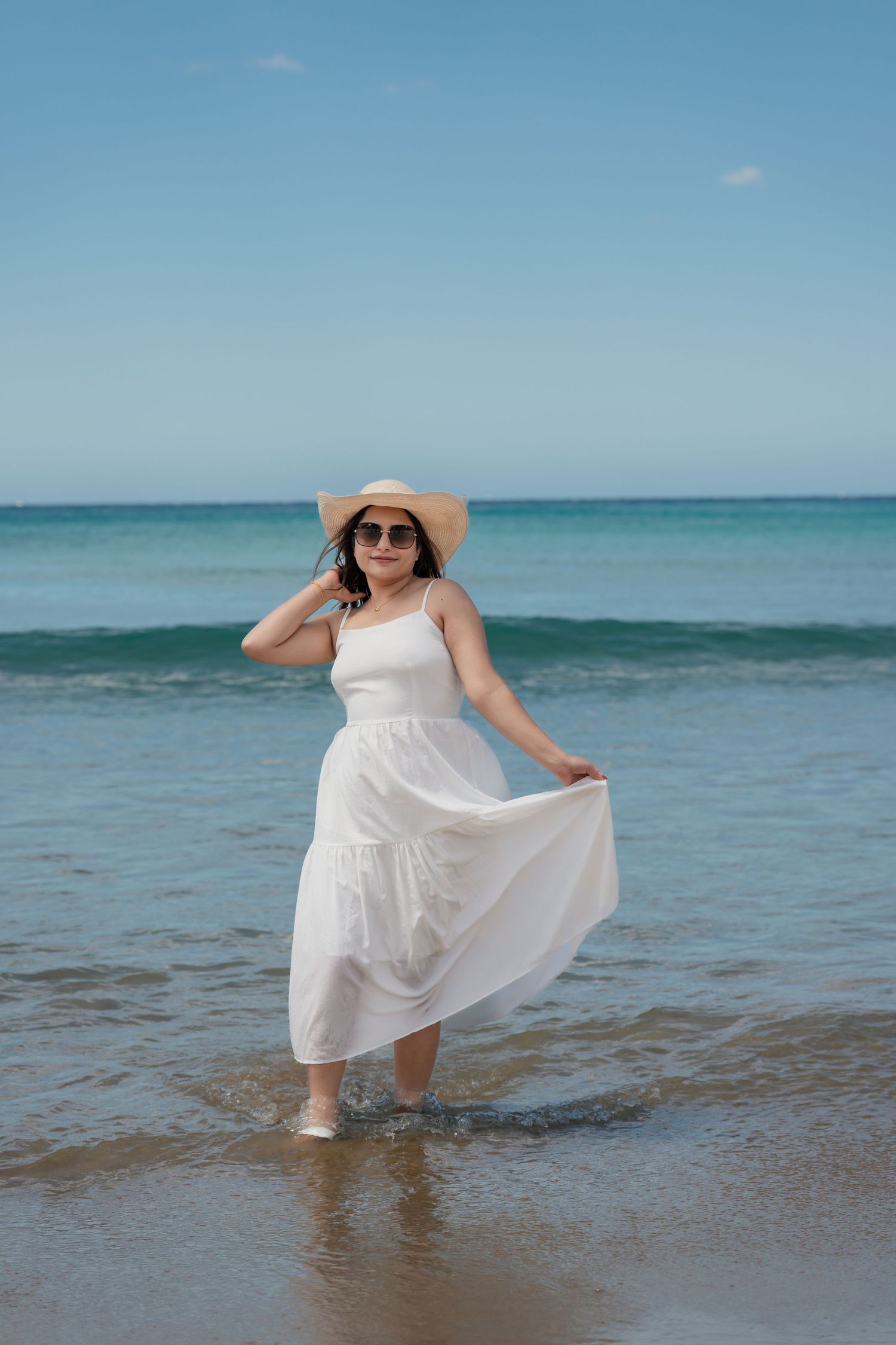 Woman in white dress and sunhat twirls playfully in turquoise ocean waters on a sunny beach day.