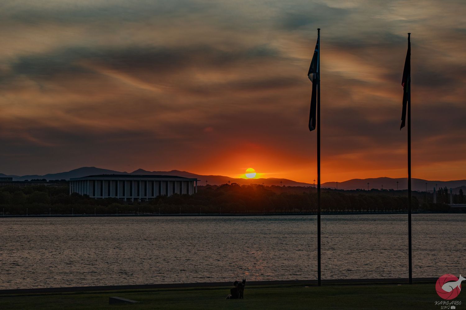 Panoramic coastal sunset with silhouetted flag poles and buildings against an orange sky.