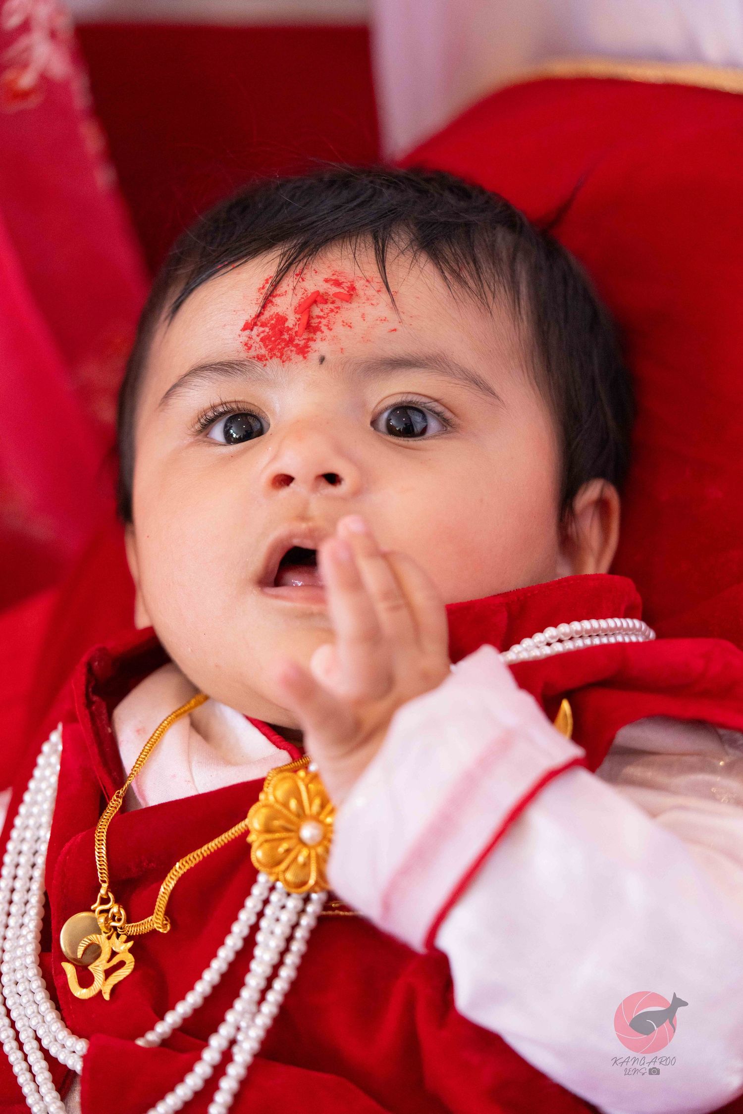 Baby wearing white outfit with traditional gold jewelry against red background.