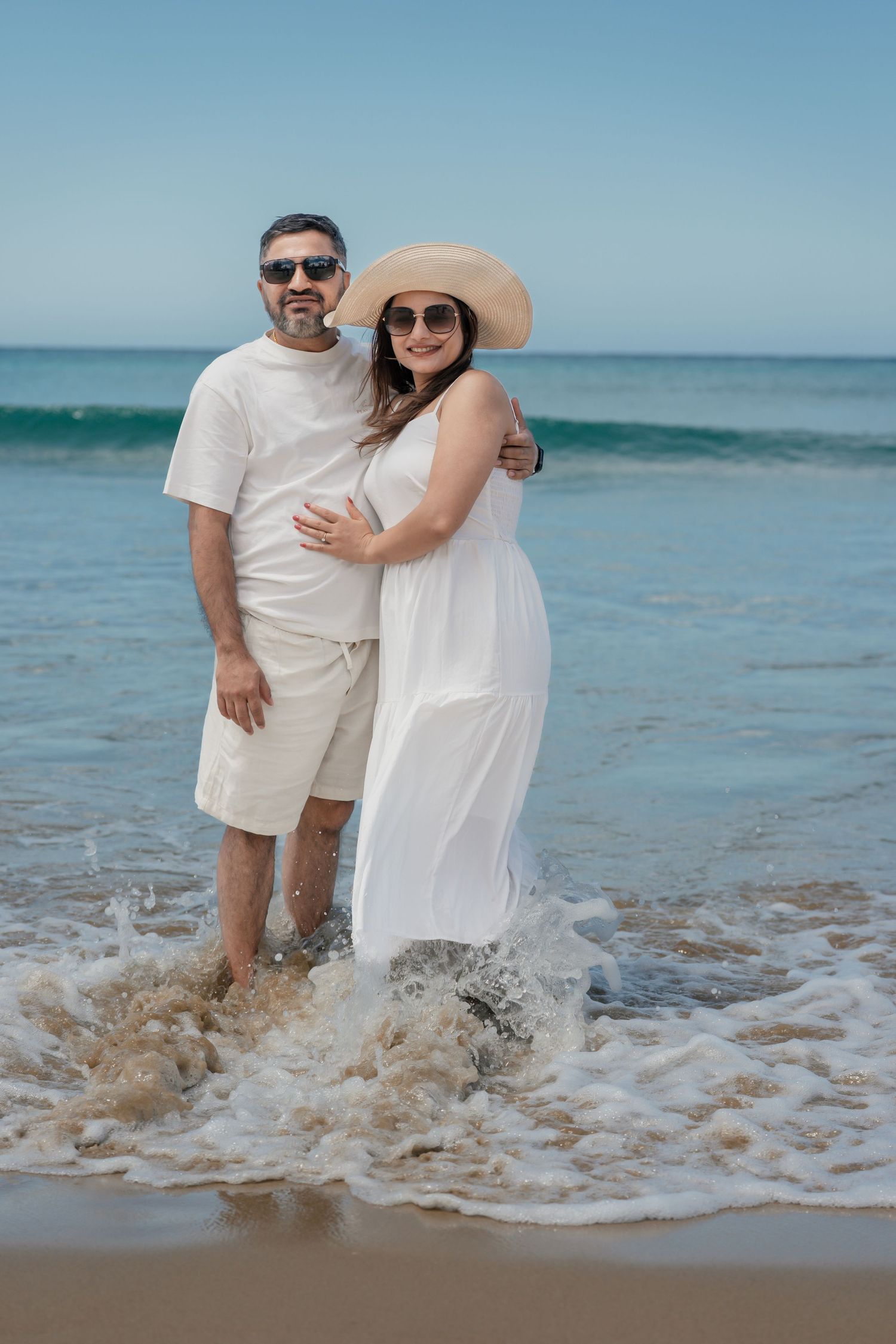 Couple wearing white outfits embraces while standing in gentle ocean waves on a tropical beach.