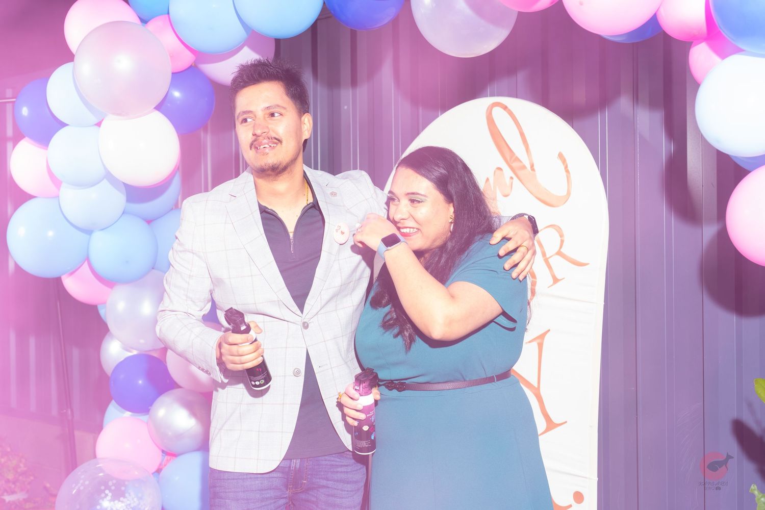 Couple celebrates at party with pink and blue balloon arch in background.