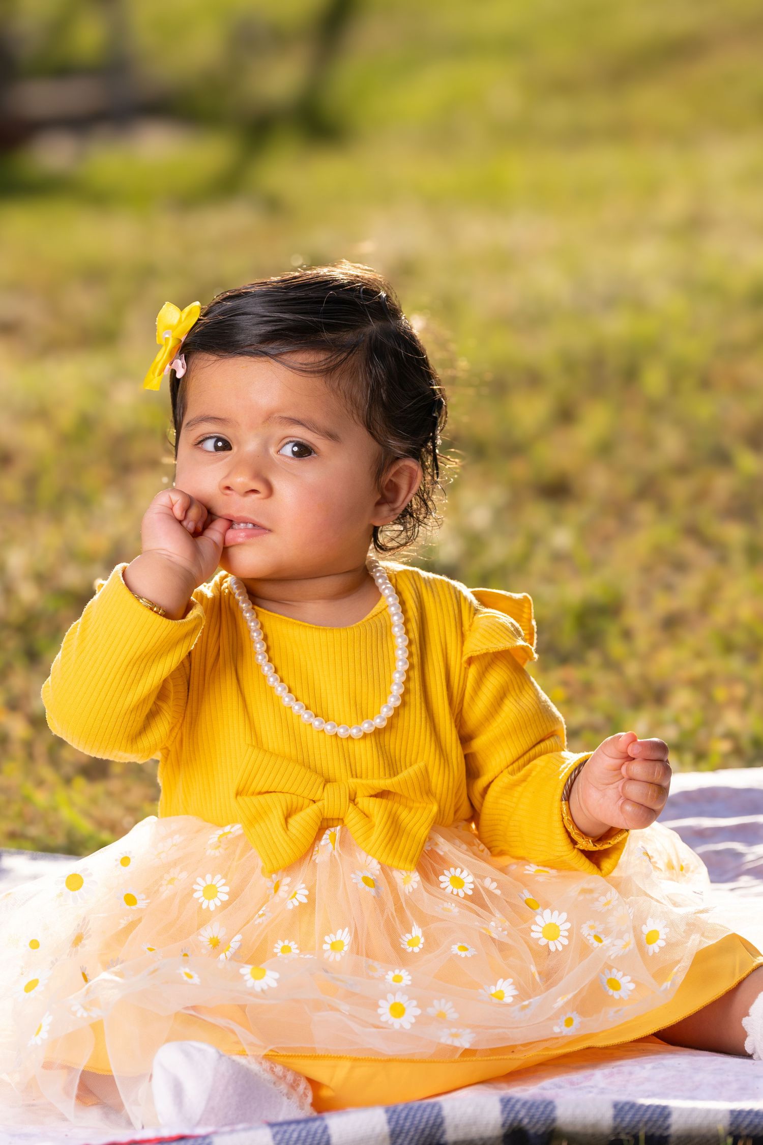 A toddler in a yellow dress with floral tulle sits on a blanket outdoors.