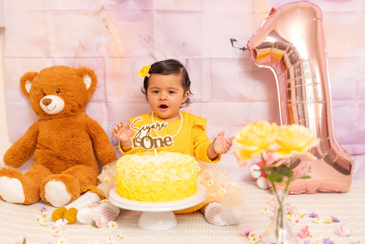 First birthday celebration setup with yellow cake, teddy bear, and number one balloon decoration.