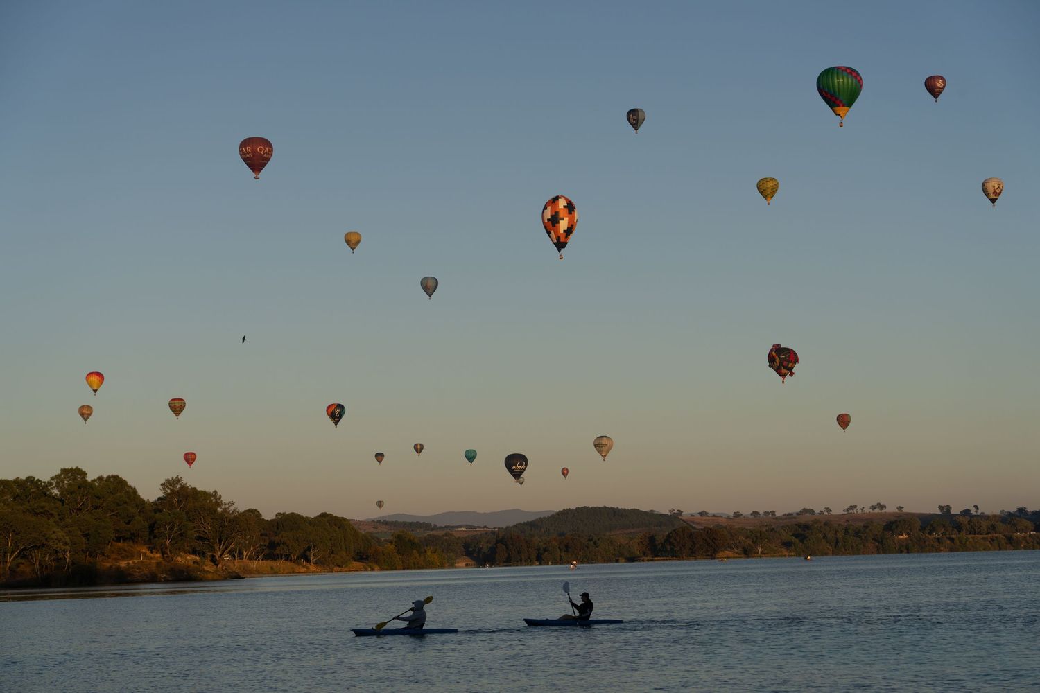 Colorful hot air balloons floating in clear blue sky over water with kayakers below.