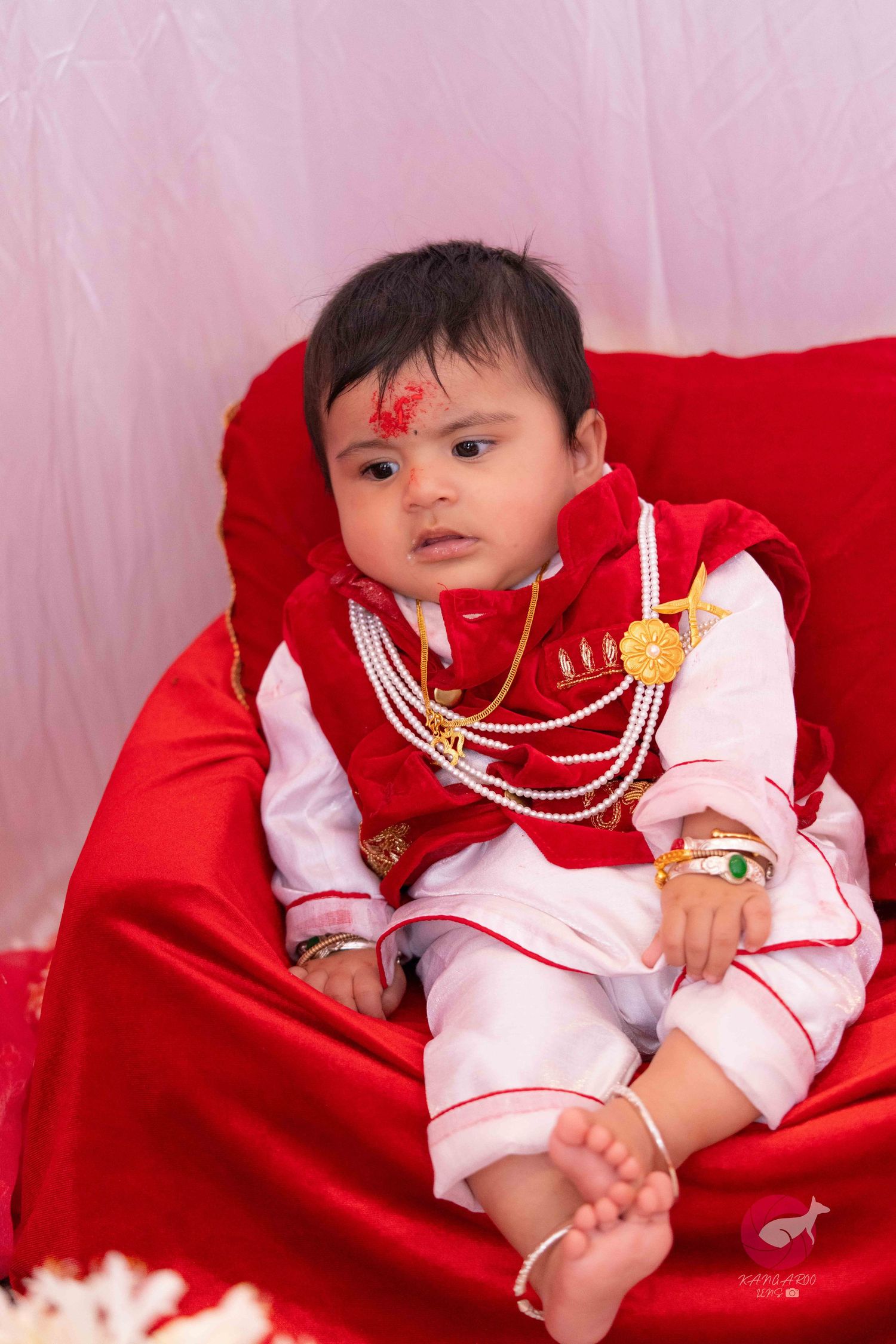 Baby in white outfit reclining on red satin fabric with traditional accessories.
