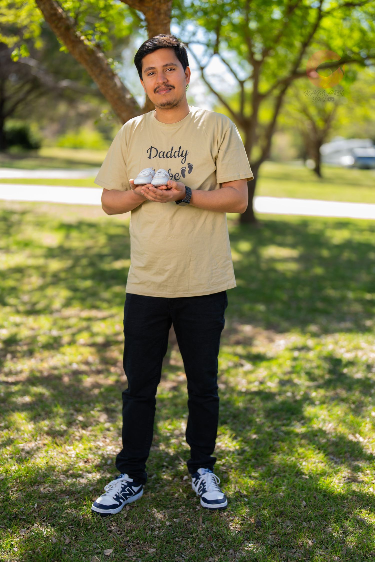Person in beige t-shirt stands outdoors in park setting with dappled sunlight.