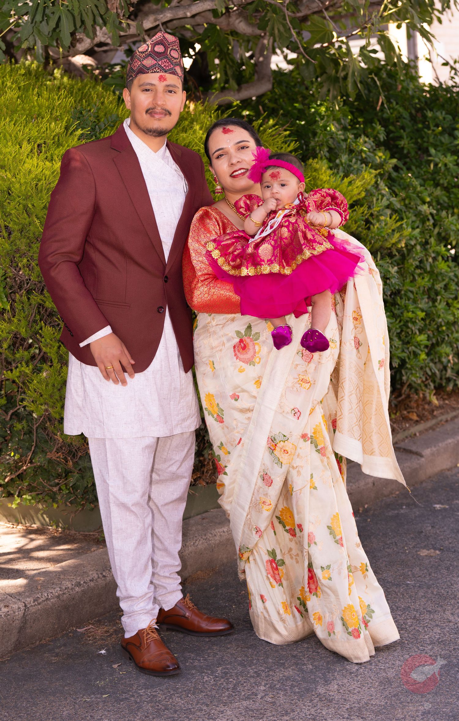 A family poses outdoors in traditional Nepali attire with a baby wearing bright pink clothing.