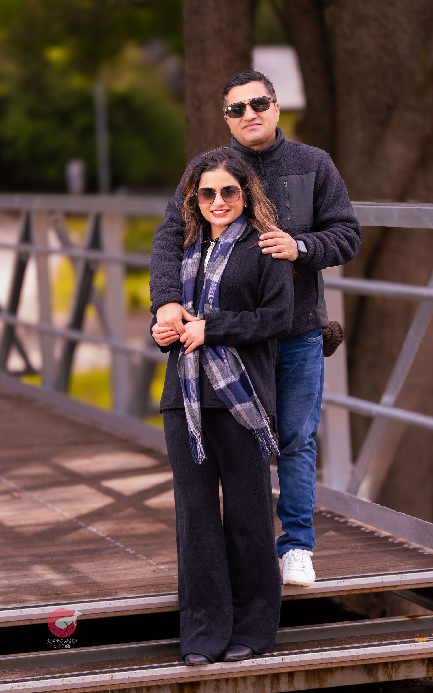 Couple in casual outfits poses playfully on wooden boardwalk surrounded by trees.