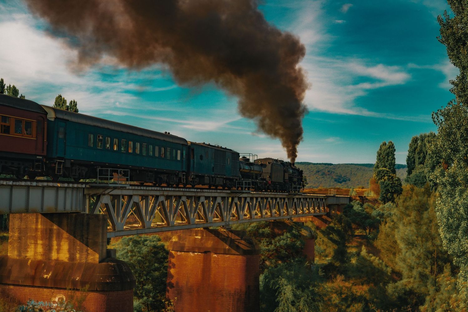Steam locomotive travels across scenic landscape with dramatic clouds and blue sky in background.