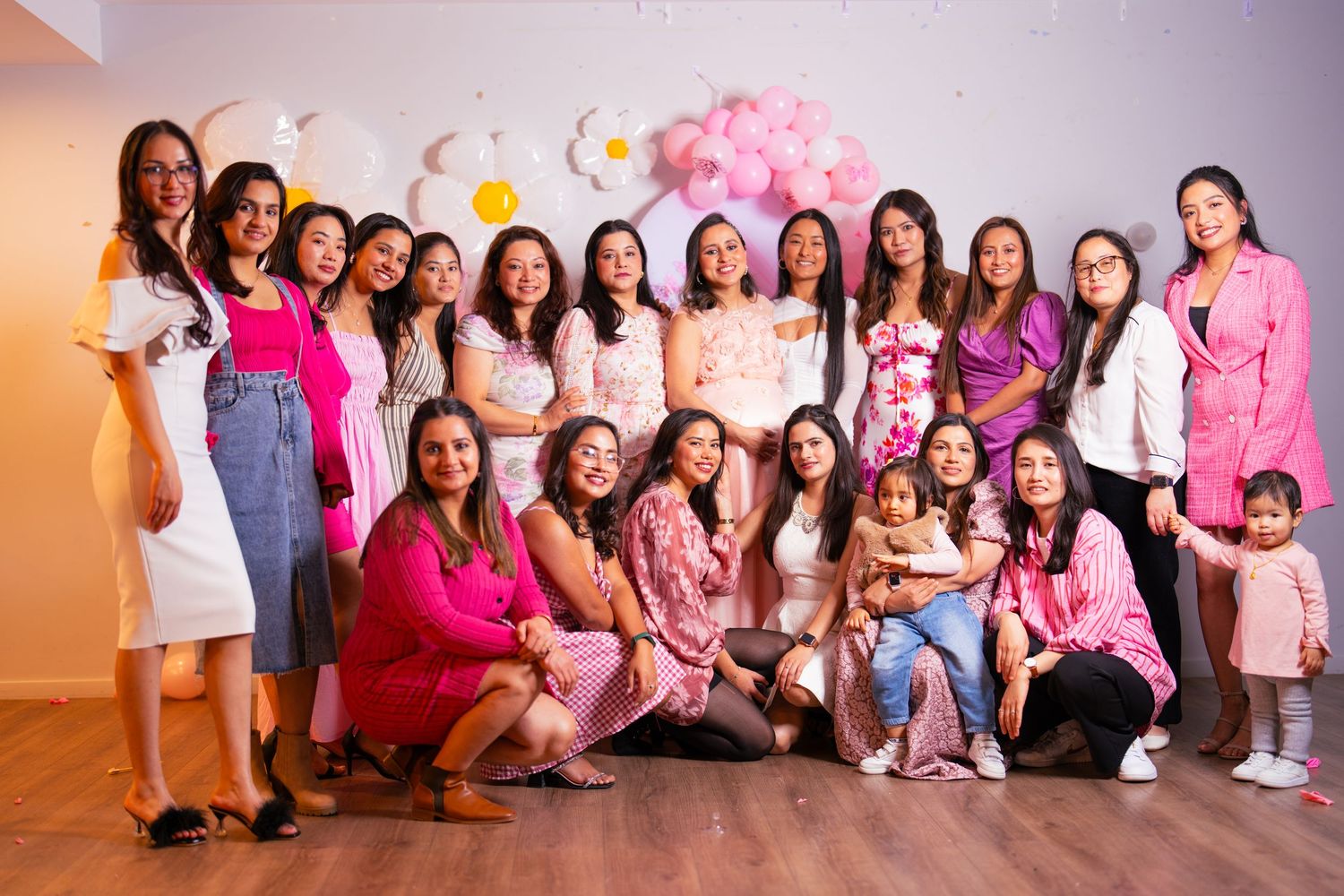 Large group wearing pink and white outfits poses together at a celebration with balloon decorations.
