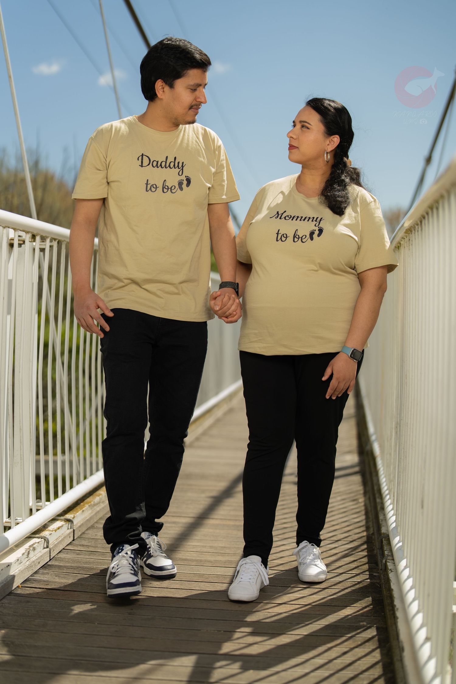 Couple wearing matching beige pregnancy announcement t-shirts walking on wooden bridge.