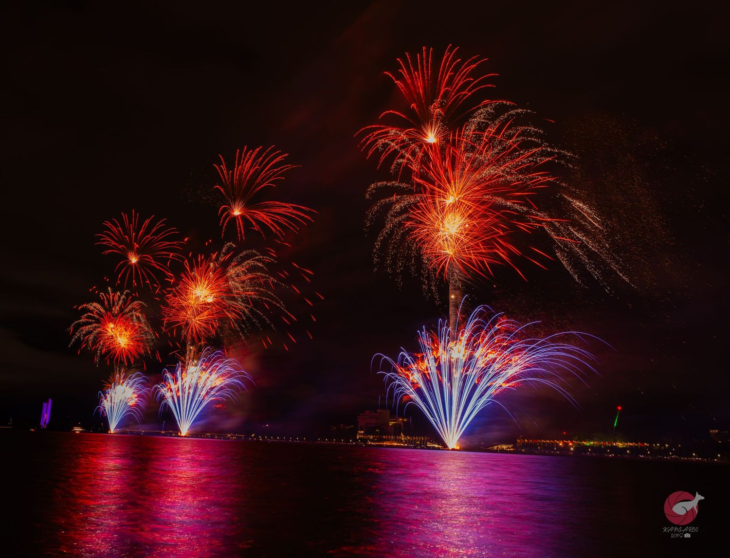 Colorful fireworks display reflecting over water at night with red and purple bursts.
