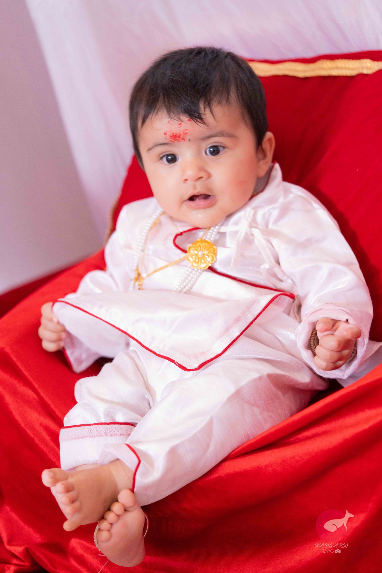Baby in white outfit lying on red satin fabric showing traditional cultural dress.