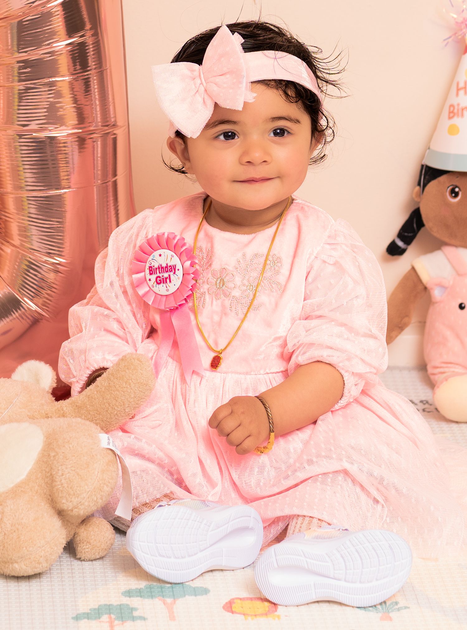 Baby in pink dress with bow headband sitting with plush toy at birthday celebration.
