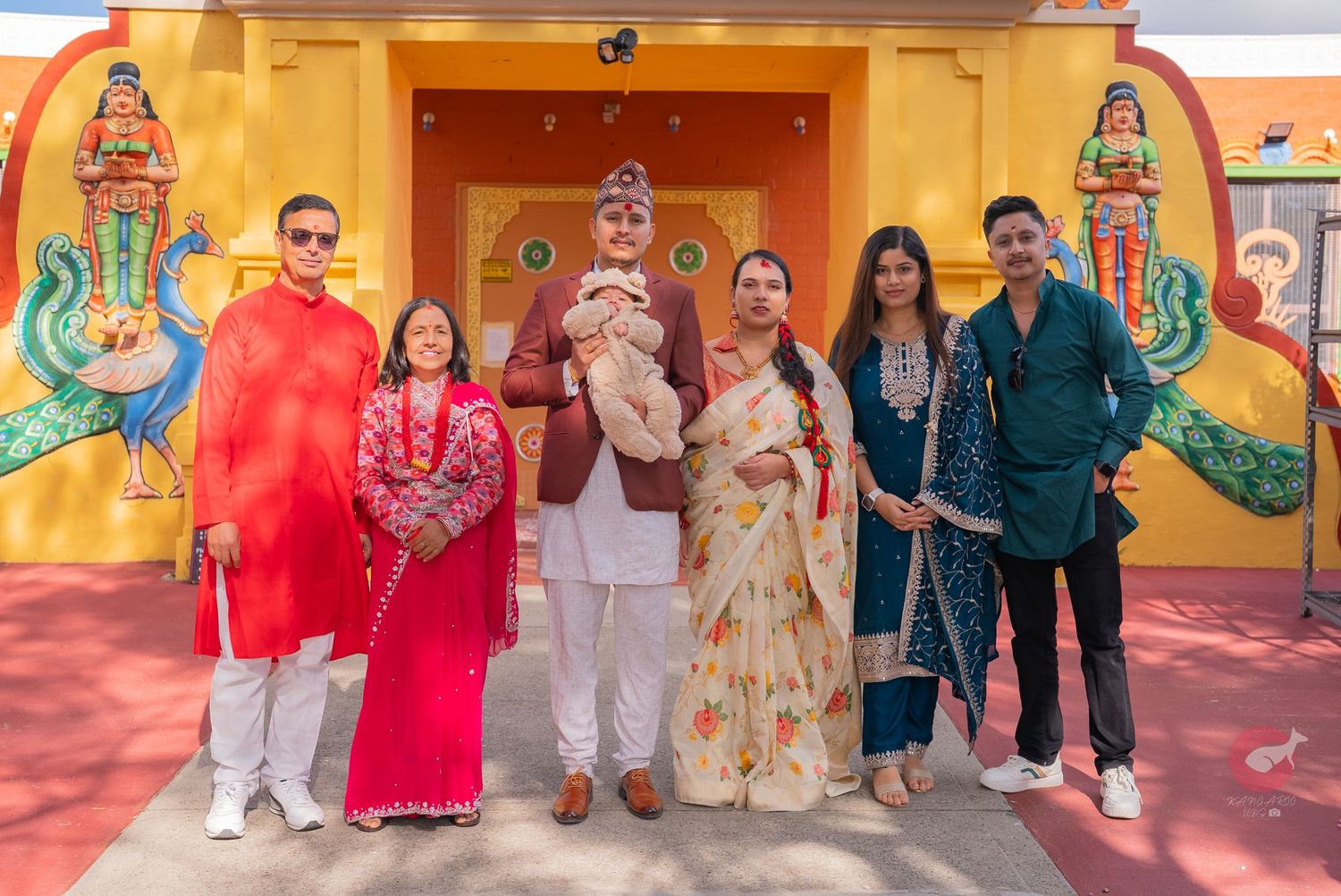 A group in traditional South Asian attire poses in front of a colorful temple wall with decorative artwork.