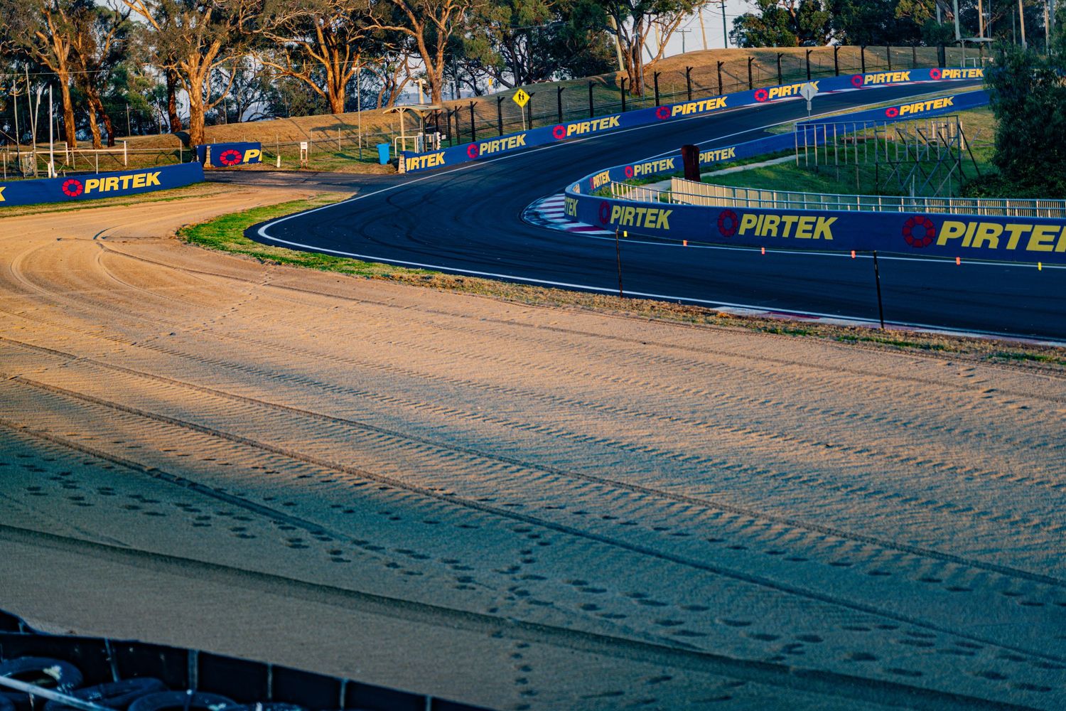 Racing circuit with blue barriers and orange runoff areas under sunny conditions.