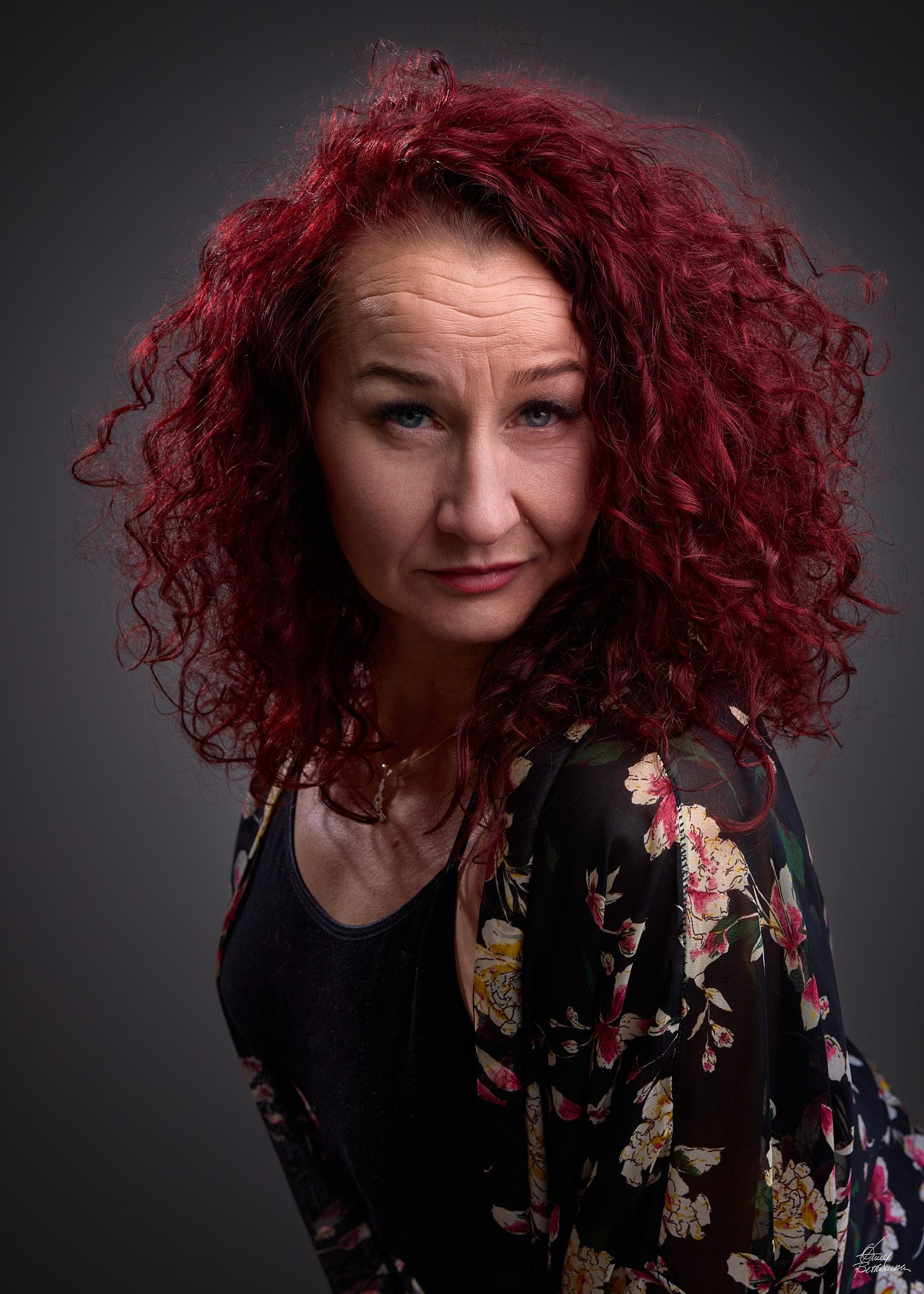 A woman with vibrant red curly hair poses expressively against a dark background in a floral patterned top.