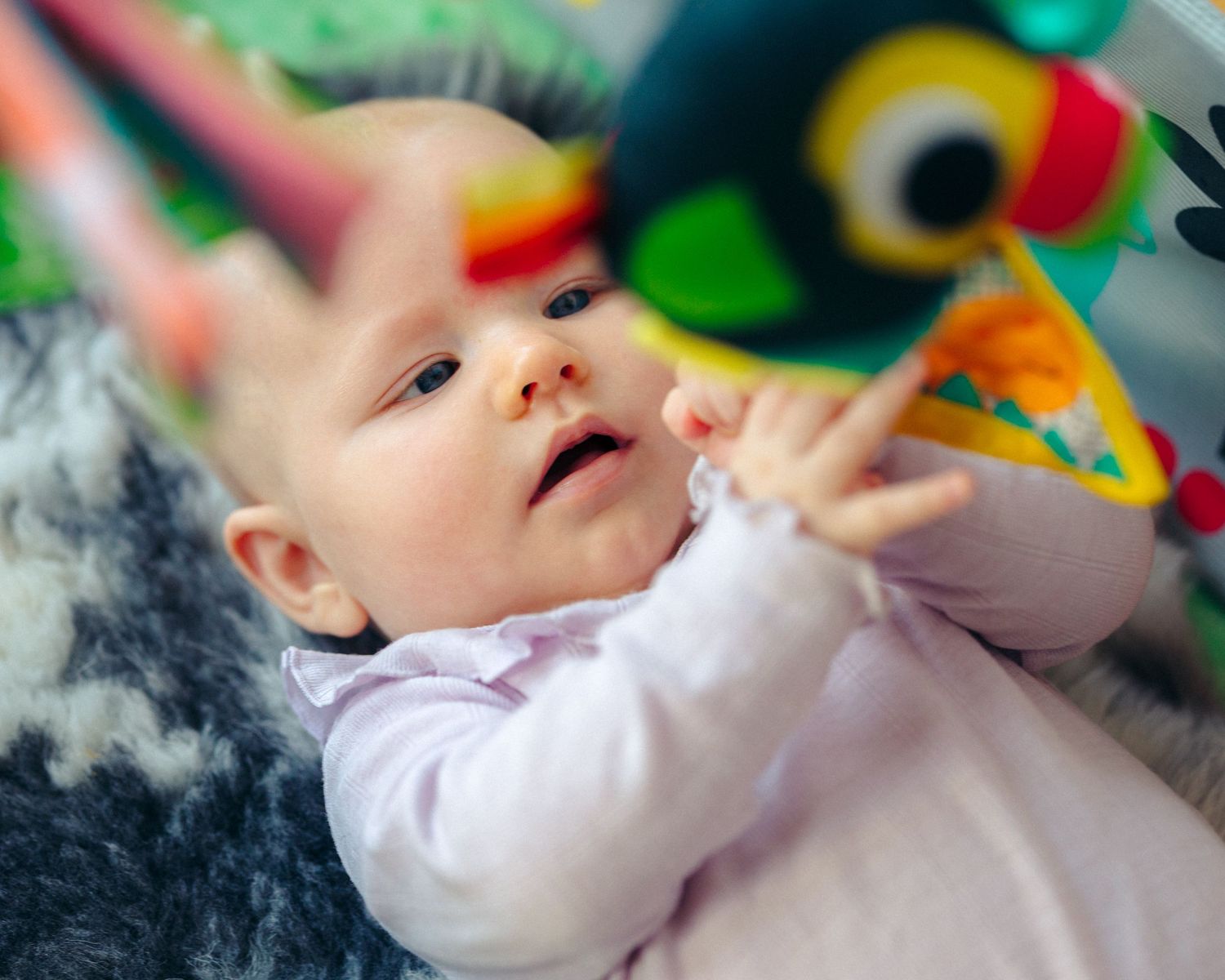 Curious baby in a light pink top lies on a soft rug, reaching toward a colorful dangling bird toy.