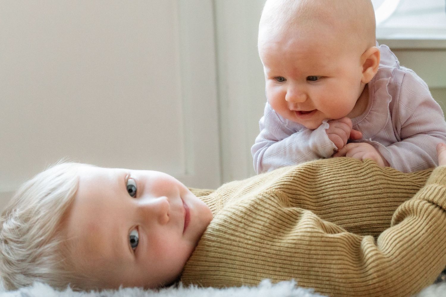 Two babies on a soft white blanket, one with blond hair lying on its back and another in a pink top smiling nearby.
