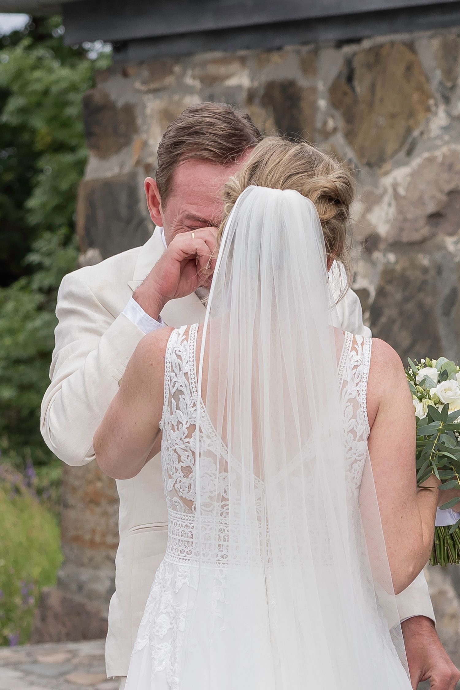 Emotional wedding first look sequence with couple in white attire by stone wall.