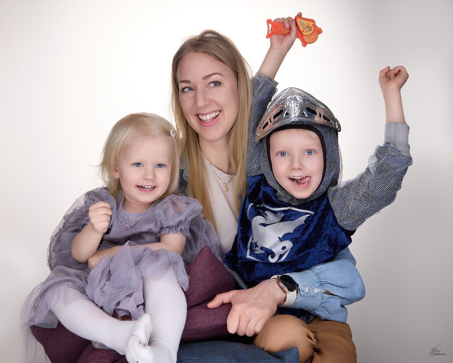 Smiling woman holding a toddler girl in a gray tulle dress and a boy in a toy knight helmet and tunic during a playful family portrait.