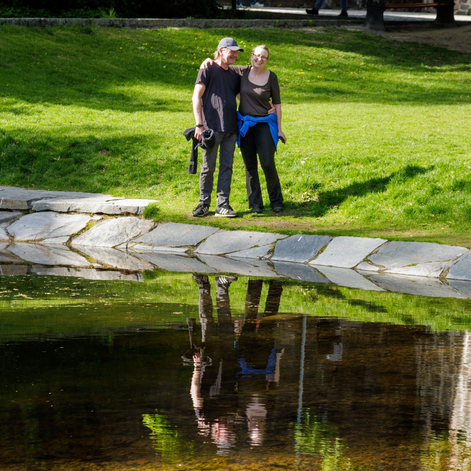 Reflections of people standing near a tranquil pond in a lush green park setting.