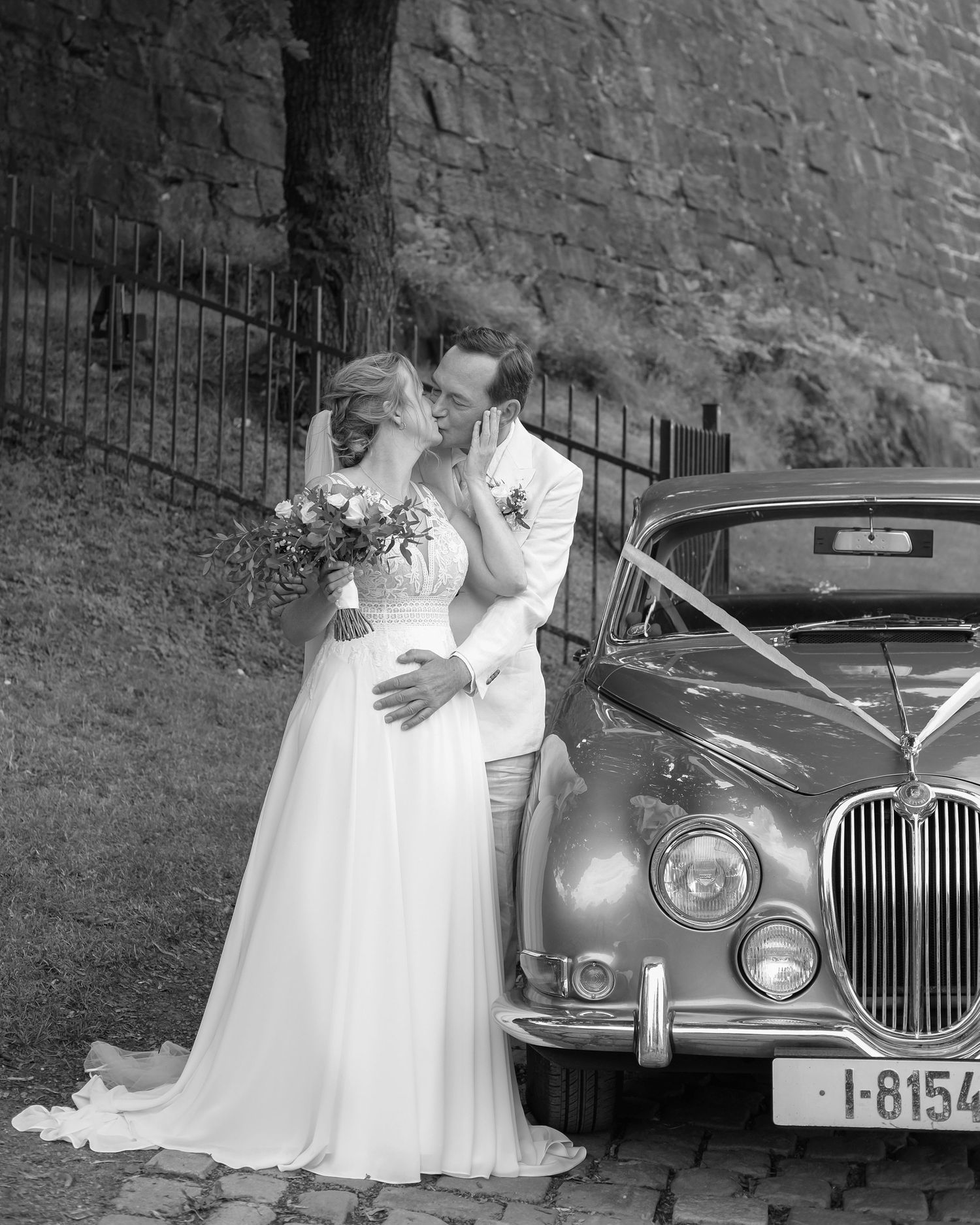 Vintage wedding photo with couple in white dress and suit posing beside classic car.