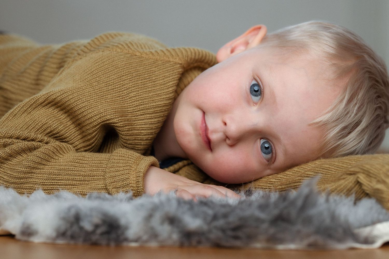 Blond toddler with blue eyes in a mustard knit sweater lies on his side, resting his head on his arm beside a soft gray fur rug.