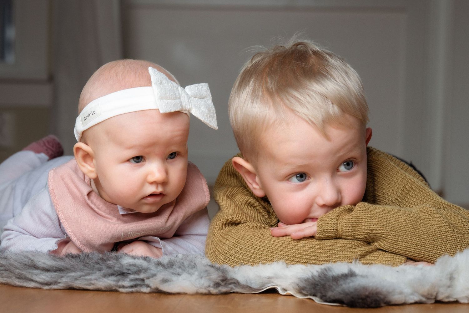 Baby in a pink top and white bow headband lies beside a toddler in an olive sweater, both resting on a soft gray fur rug.