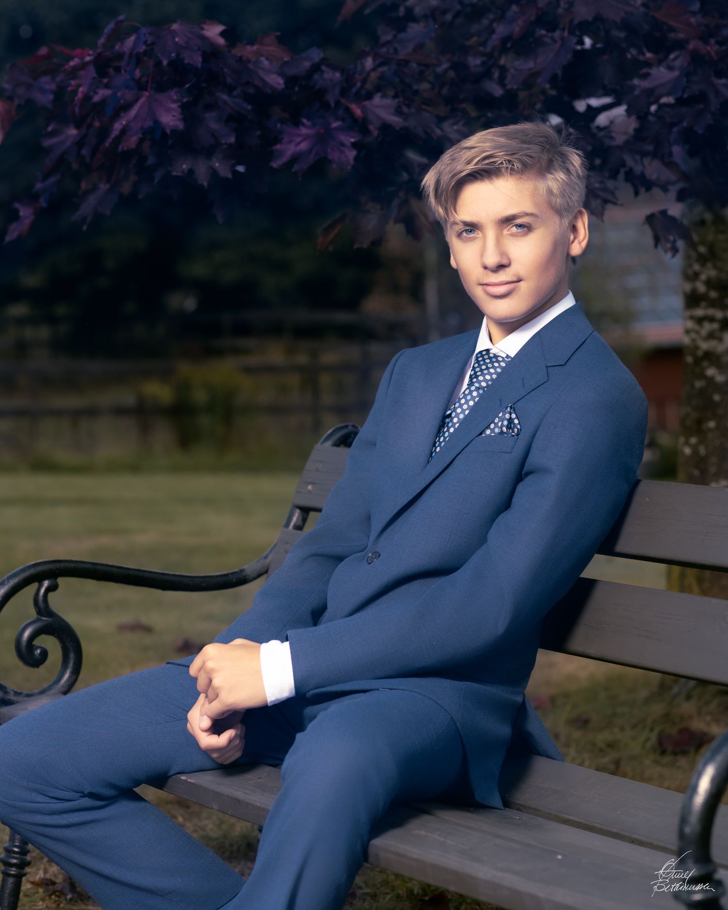 Young person in a navy blue suit sitting on a park bench with purple foliage in background.