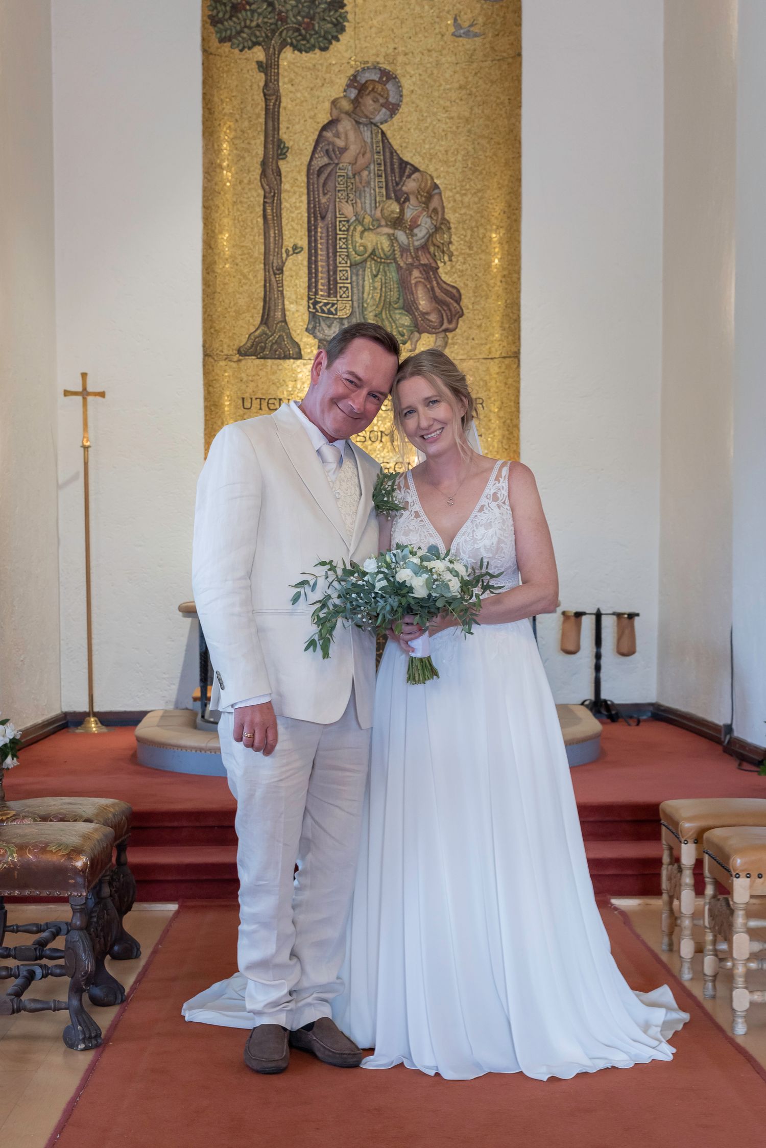 A couple in white wedding attire sharing an intimate moment in a church with golden mosaic walls.