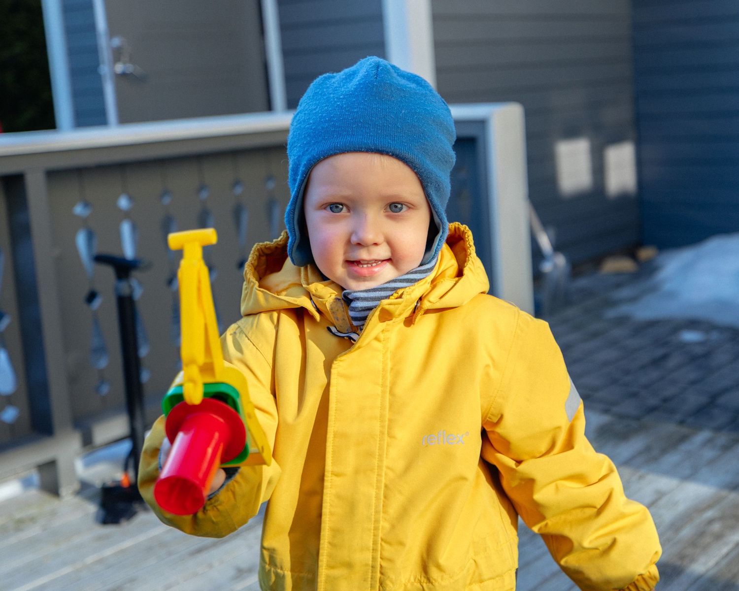 Smiling toddler in a yellow raincoat and blue knit hat holds a colorful toy tool while standing on an outdoor wooden deck.