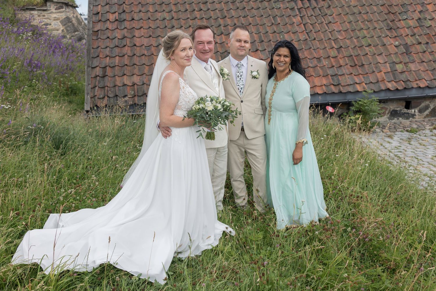 Wedding party in mint green and beige attire poses together on grassy hillside near rustic building with terracotta roof.