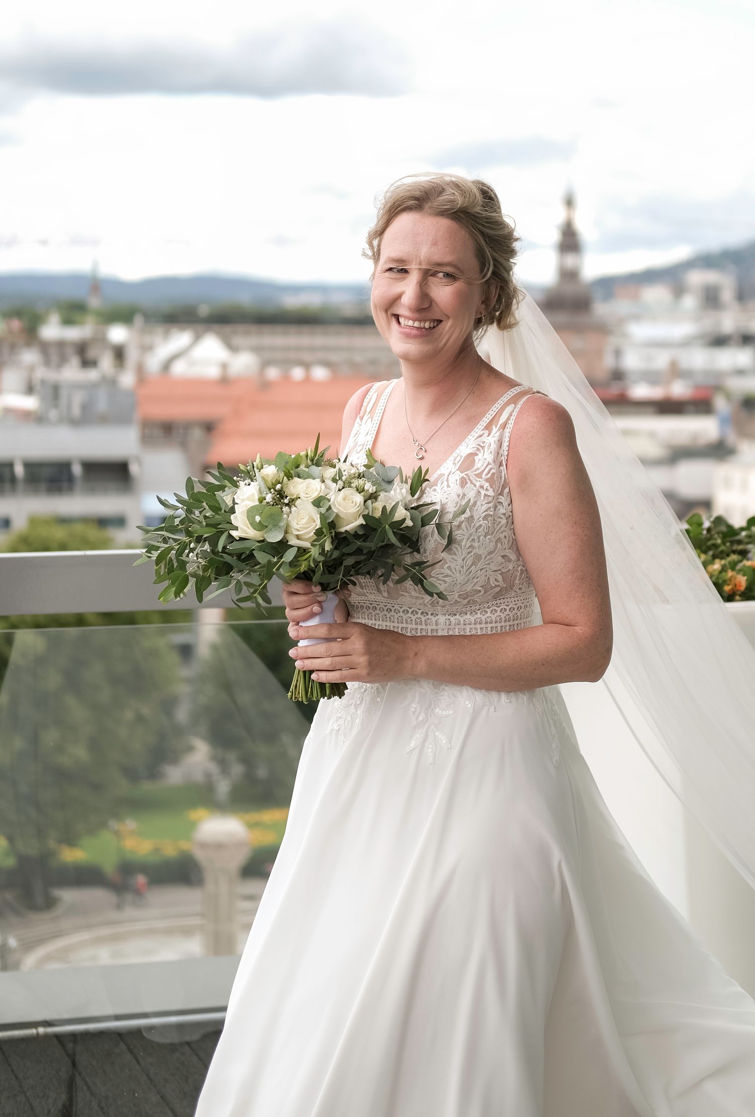A beautiful bride in a flowing white dress holds a lush green and white floral bouquet against a city backdrop.