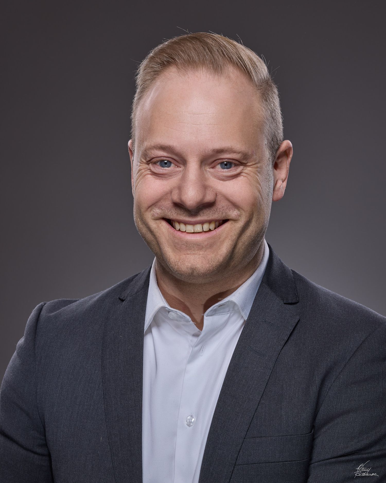 Professional headshot portrait of someone in a dark suit and white shirt against a gray studio background.