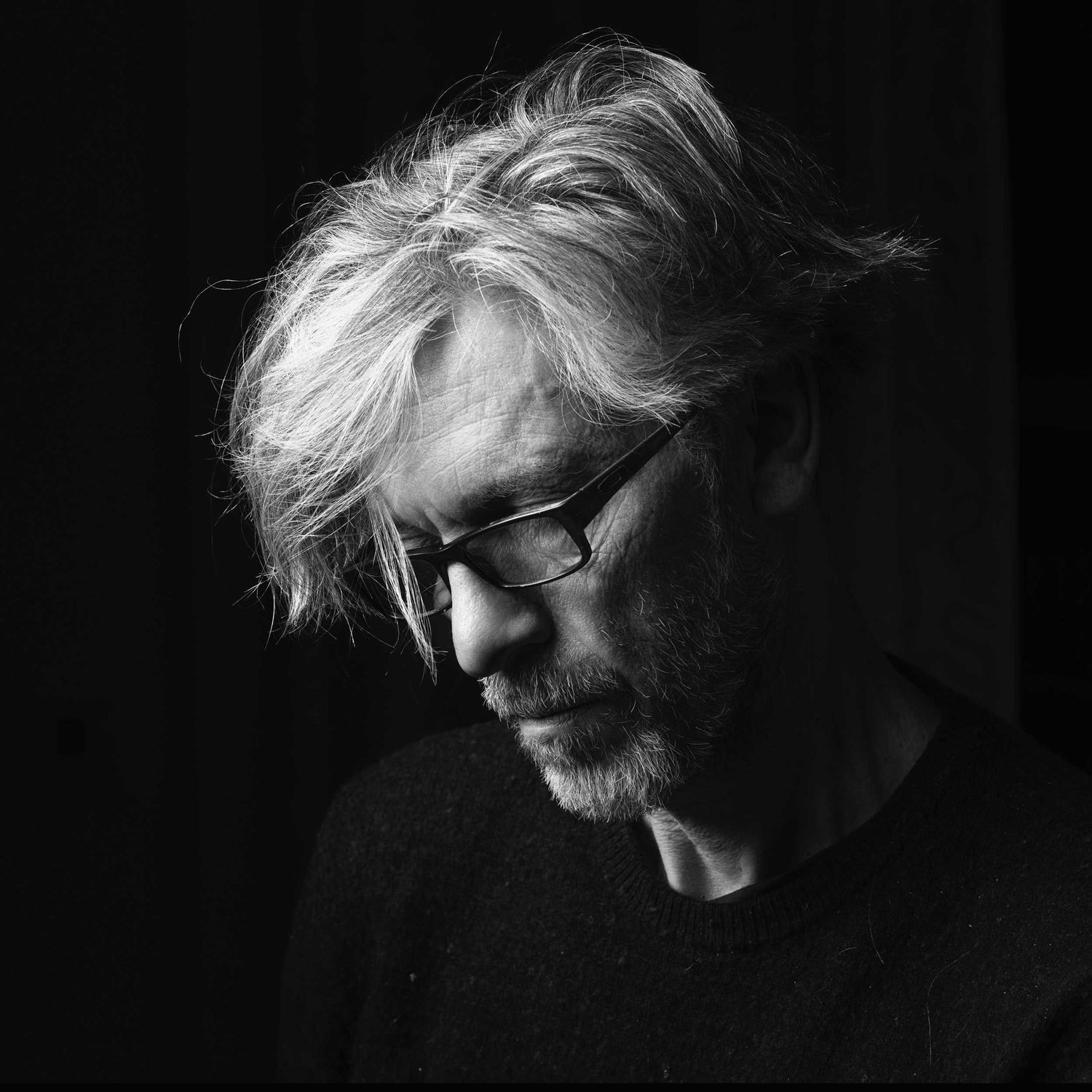 Dramatic black and white portrait of person with tousled gray hair and glasses in dark lighting.