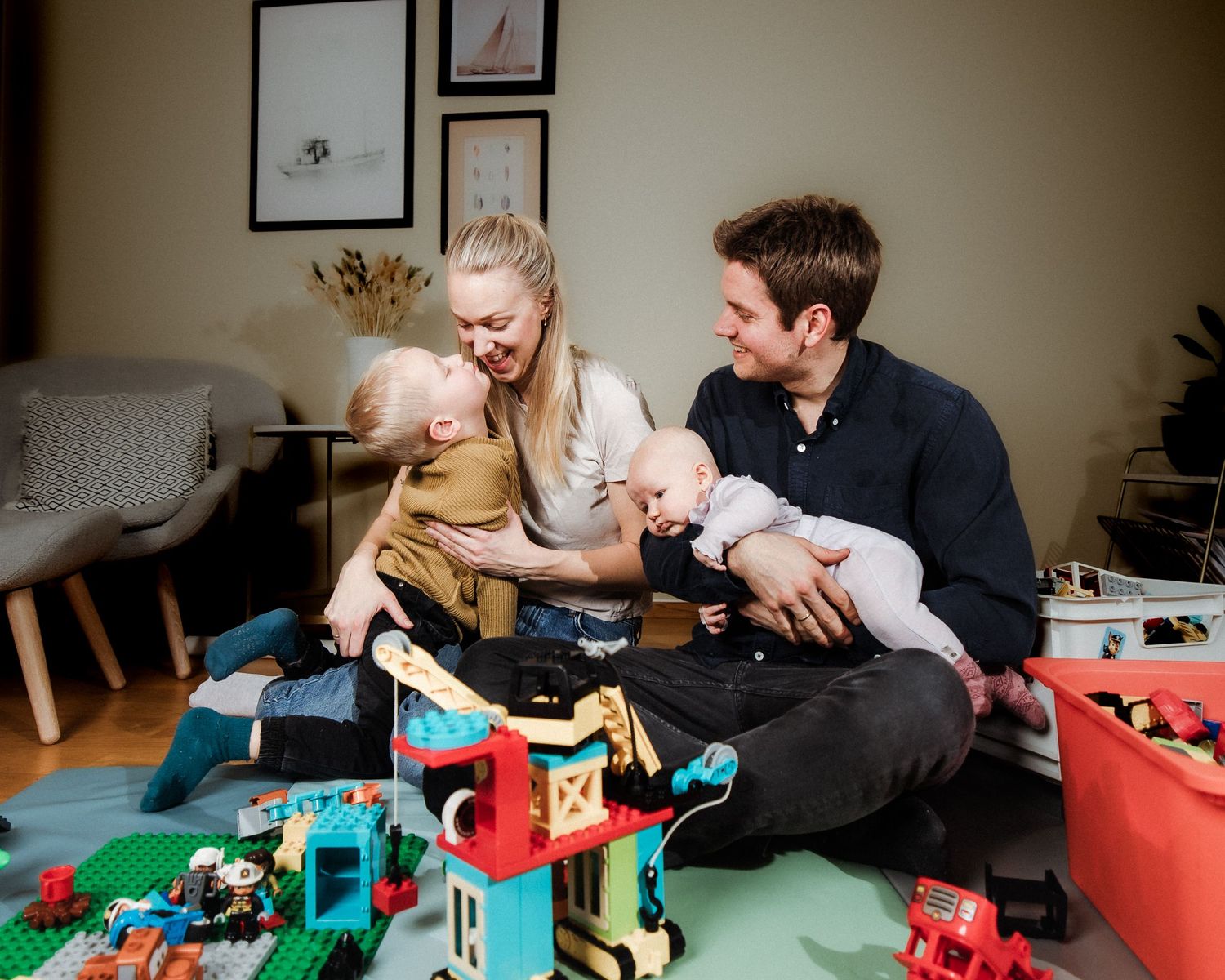 Smiling parents sit on a playroom floor with a toddler and a baby, surrounded by colorful building blocks and toys in a cozy living room.